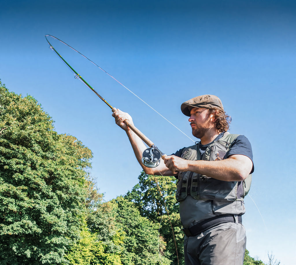 A man wearing a cap and a gray fly-fishing vest stands outdoors under a clear blue sky, holding a bent fly fishing rod with a green line. Lush green trees fill the background on the left.